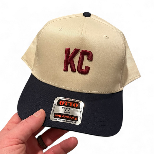 Beige and navy baseball cap with 'KC' logo held by a hand on a white background