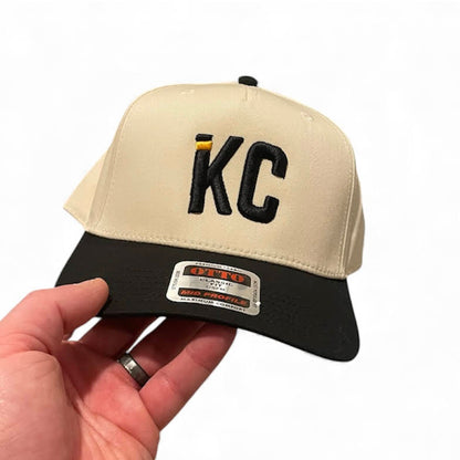 Beige and black cap with 'KC' logo held by a hand on a white background